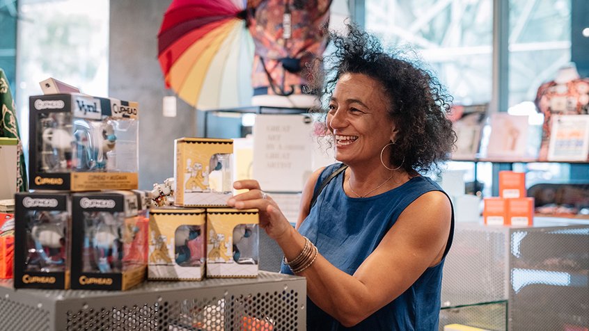 A woman browses items in the ACMI Shop. She is wearing a blue top and smiling. Photograph by Phoebe Powell