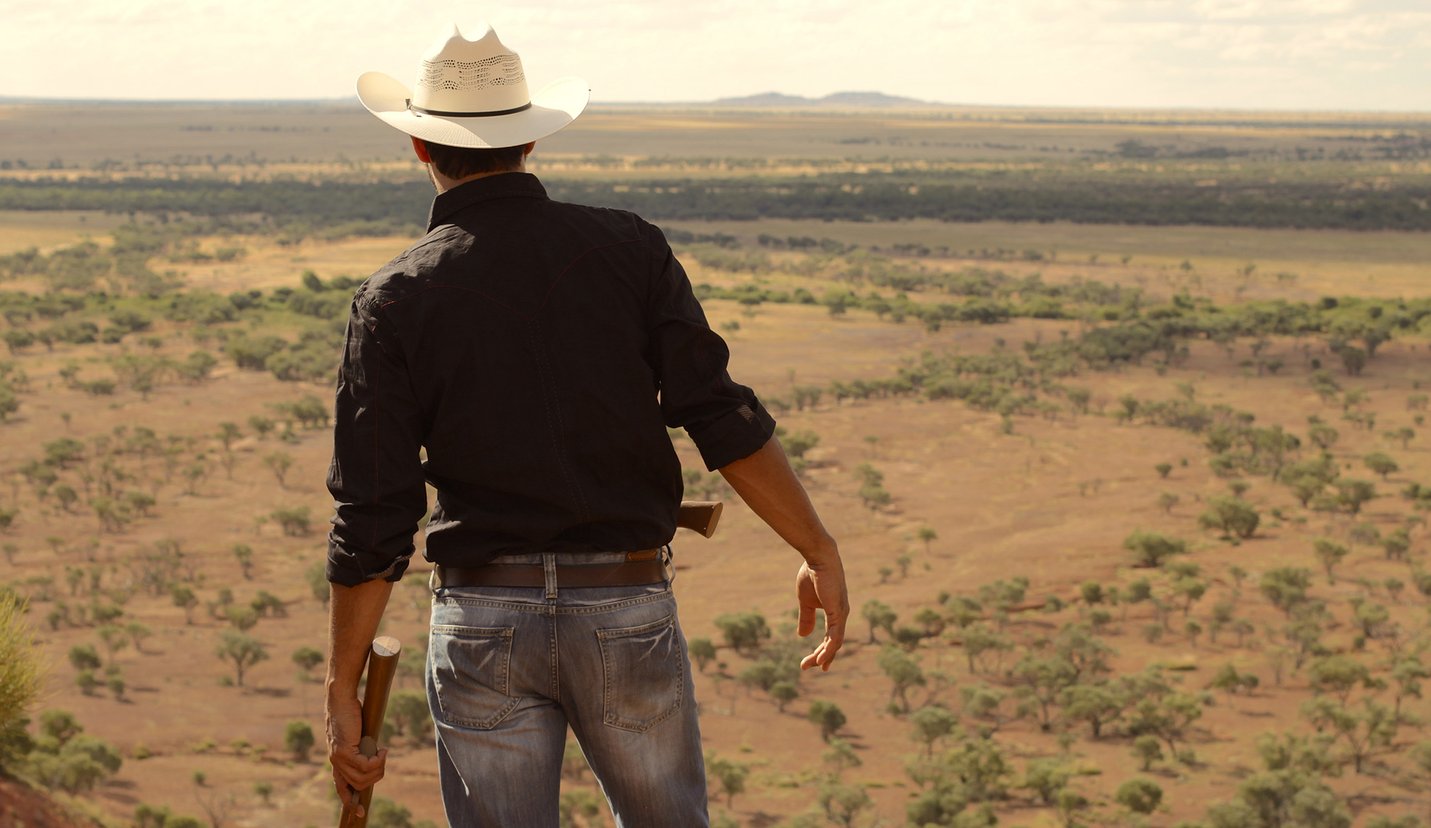 A rear shot of a man in Australian outback attire surveying the landscape from a high elevation