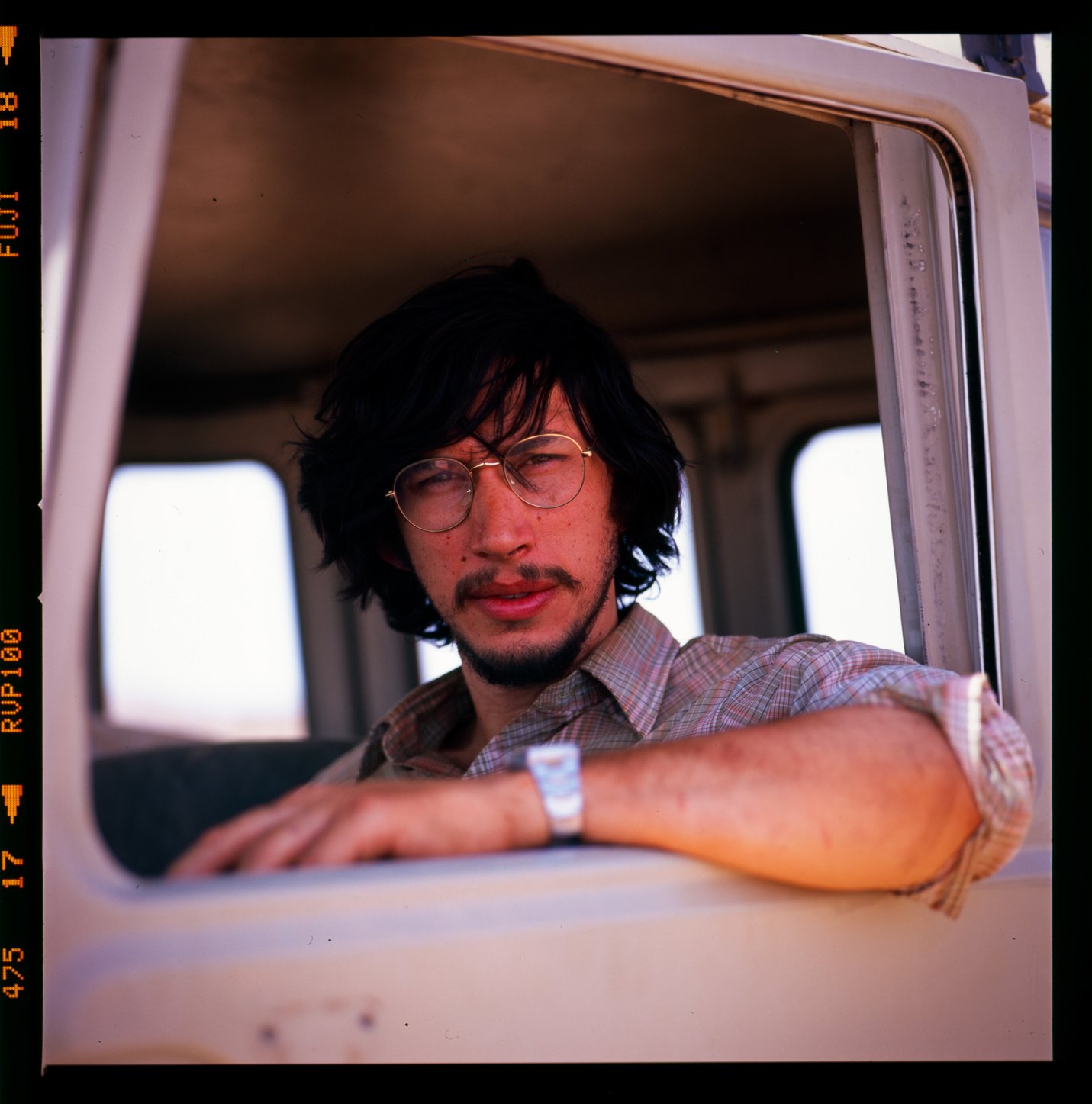 Adam Driver on the set of the movie Tracks. He is wearing glasses and looking into the camera while sitting in a car and resting his elbow on the ledge of the car window.