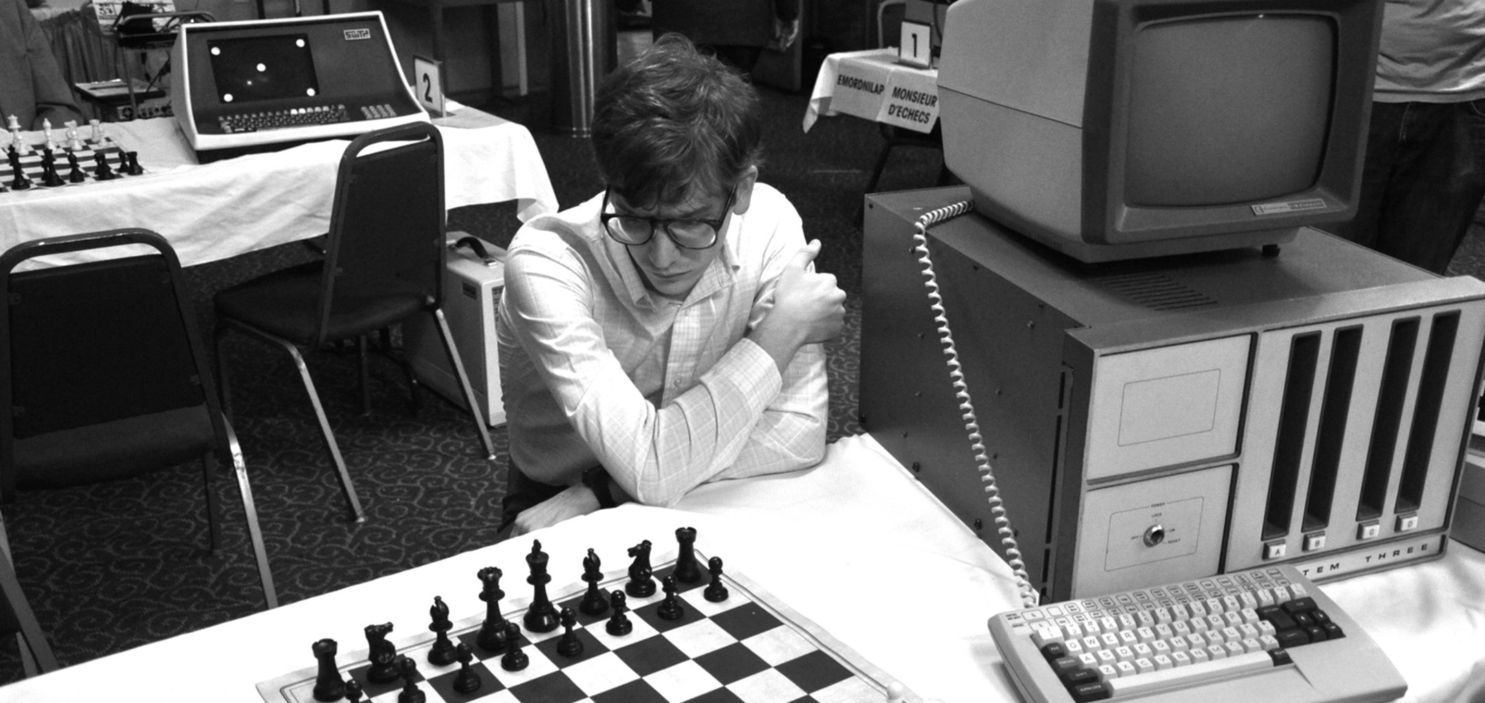 Black and white image of man in thick glasses staring down at a chess board. He is surrounded by 1980s computers.