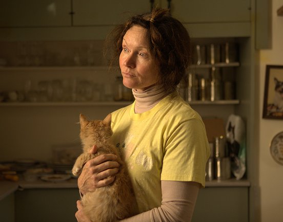 A middle-aged woman (played by Essie Davis) in a yellow top stares out pensively to the left of the frame, while holding a ginger cat. She has short brown hair and is standing in a kitchen.