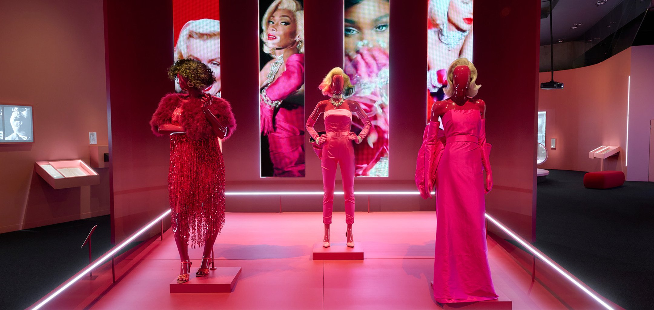 3 Mannequins in pink outfits standing on a pink plinth, with moving images of Winnie Harlow and Marilyn Monroe in the background