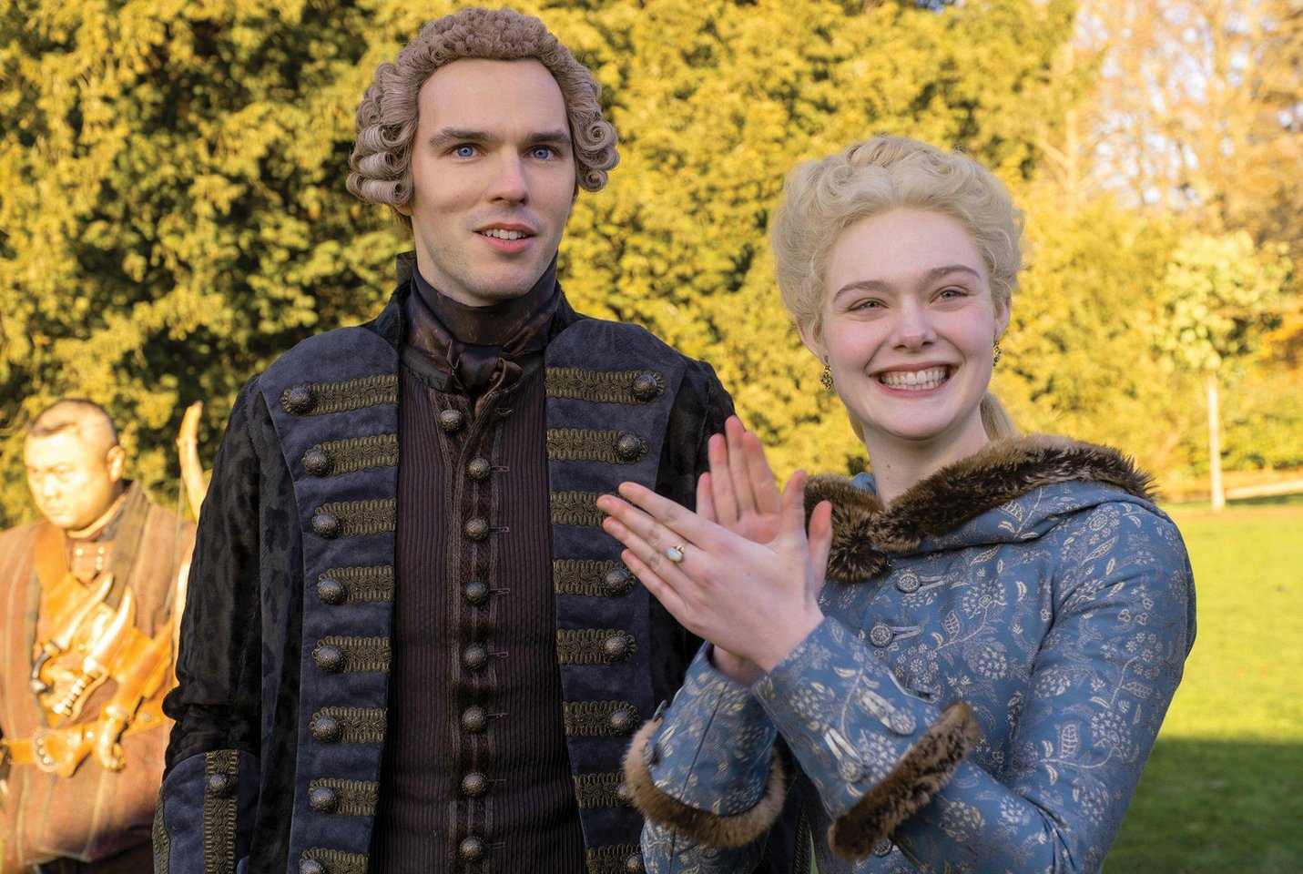 Nicholas Hoult and Elle Fanning as Peter and Catherine, standing in a field wearing regal, period outfits. Fanning is applauding with a big smile on her face.