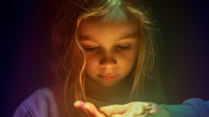 A young girl looks at films projected on her parent's hand in Memory Garden. Rainbow colours are reflected on her face.