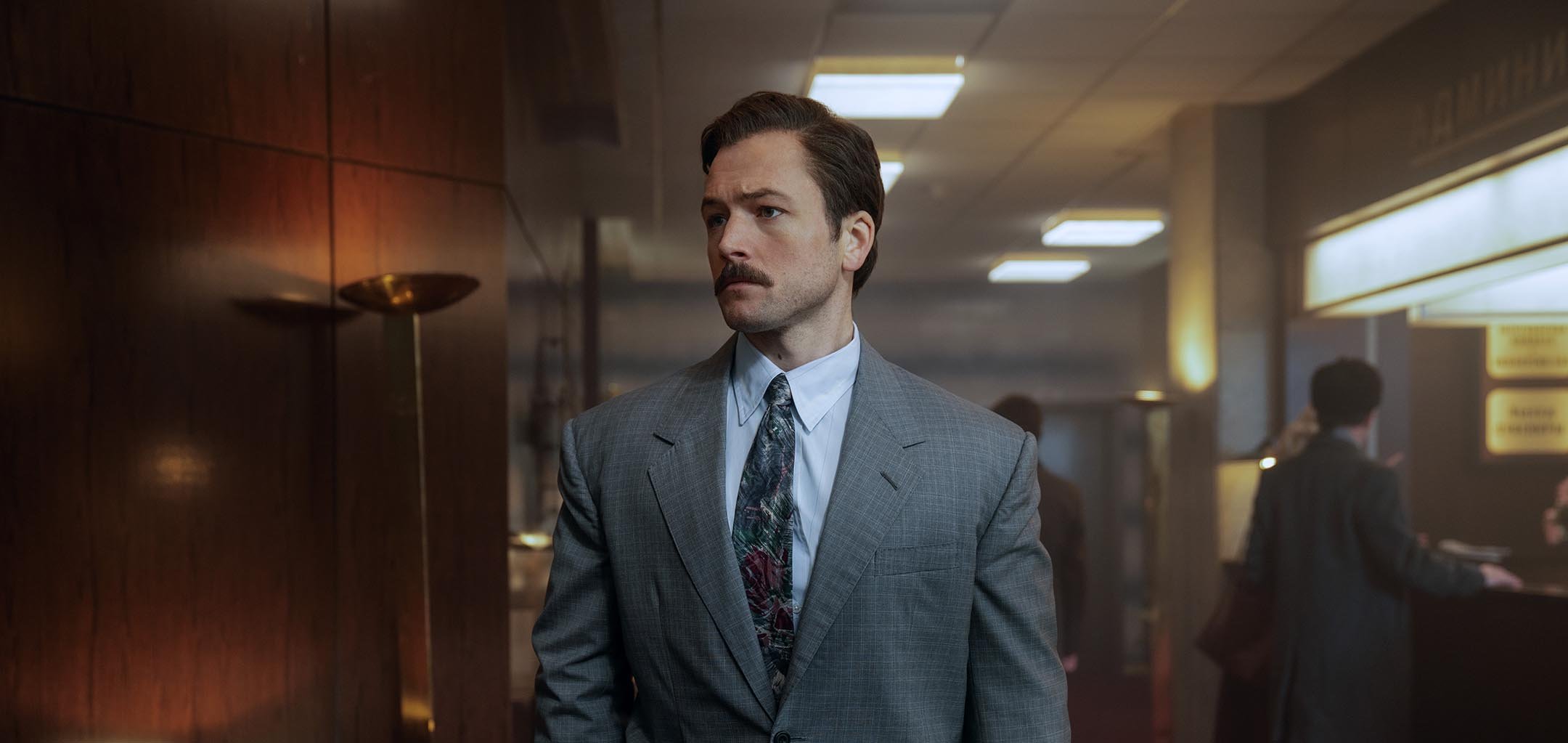 A moustached man in a grey suit (Taron Egerton) looking to the left. He is standing centre of frame, and he is in an office environment.