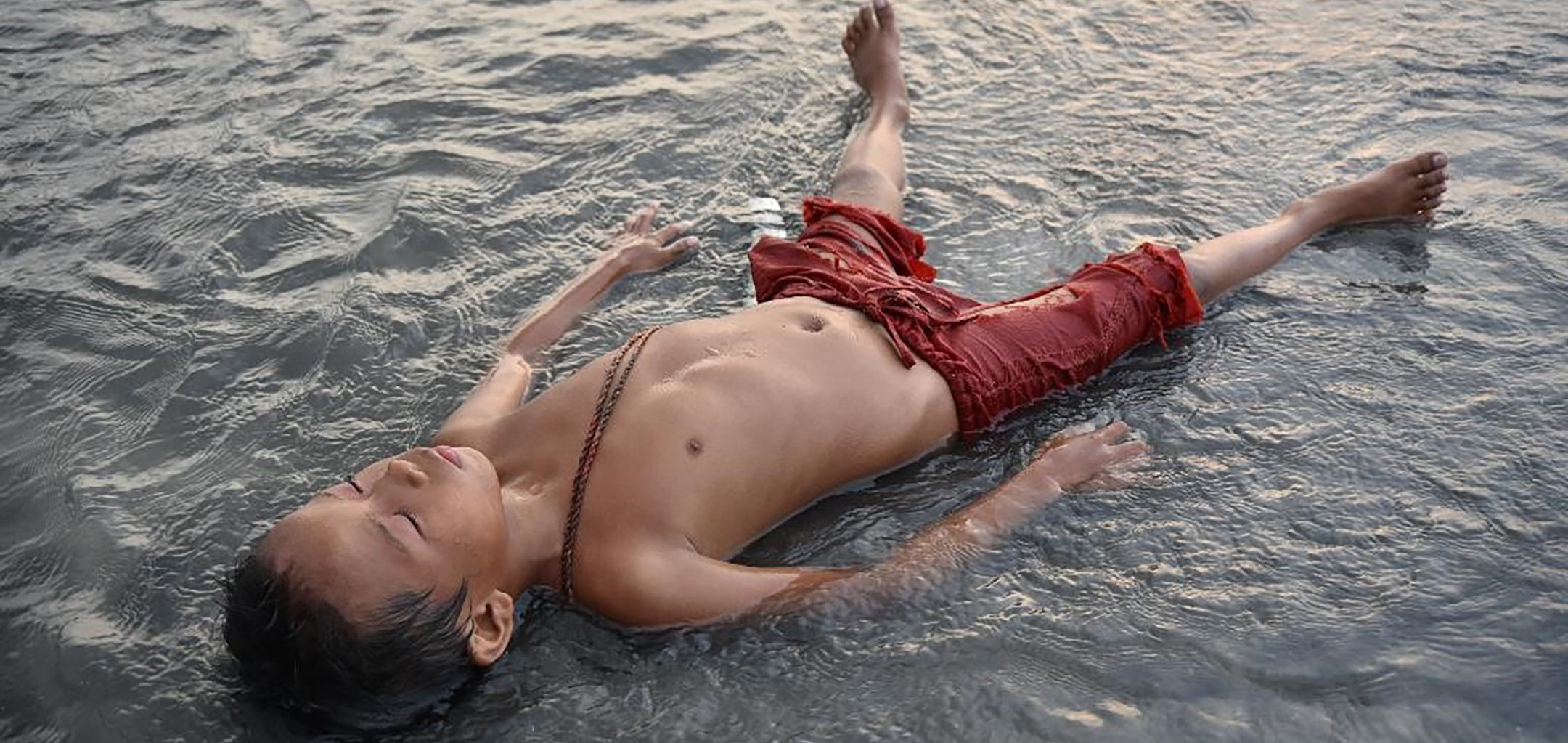 A young Filipino boy in red shorts, lying on his back with his eyes closed in a shallow body of water.