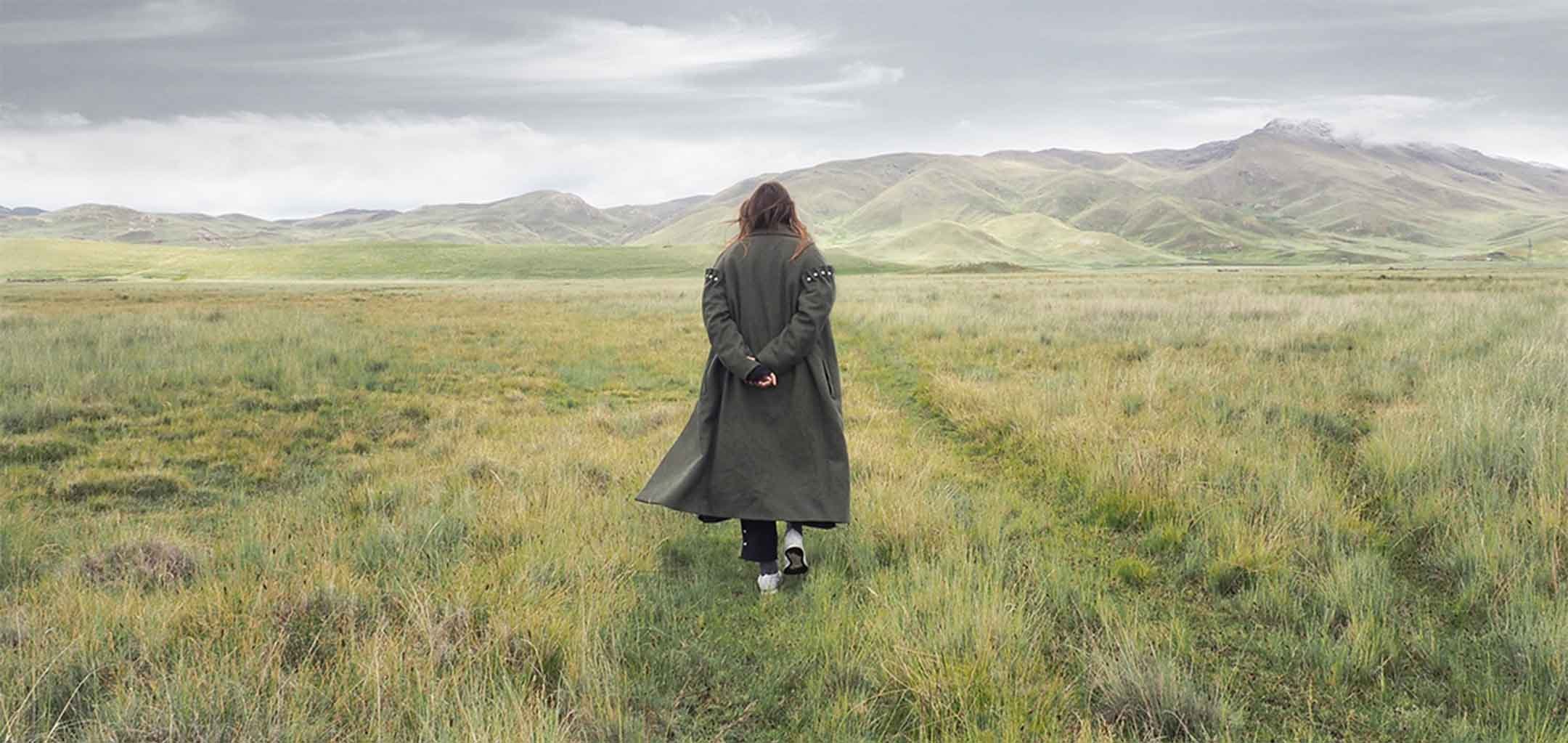 A woman in an olive-coloured trenchcoat is walking away in the centre-of-frame. She is surrounded by greenery and mountains. Her arms are folded behind her back.