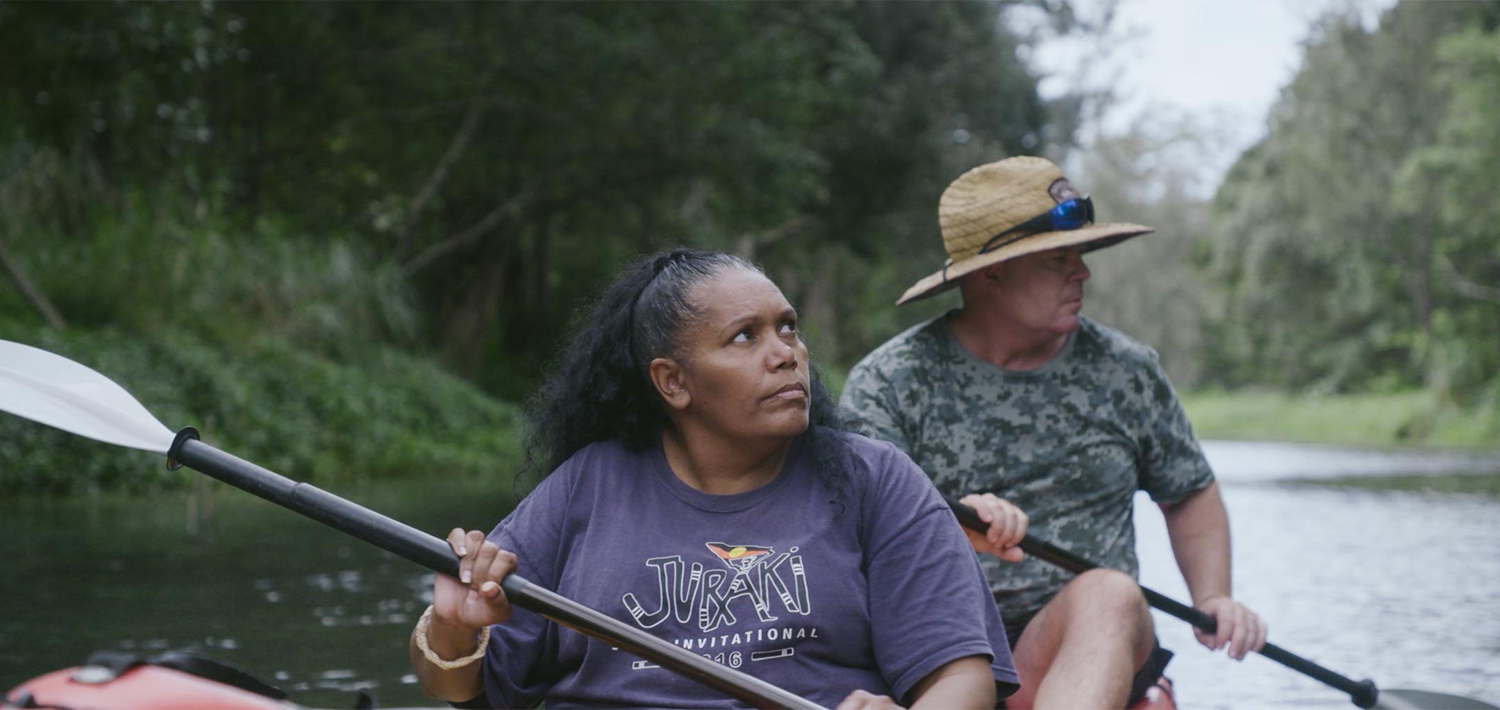 A woman is looking into the distance as she holds an oar while sitting in a boat. A man behind her in the boat is also holding an oar, and wearing a large straw hat. The boat is in a river surrounded by a green, bushy area.