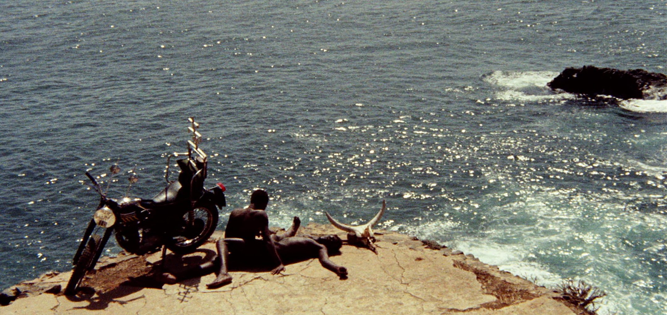 Two African men, naked, lying on a rocky platform overlooking the sea. There is a motorbike and a horned cow skull on either side of them.