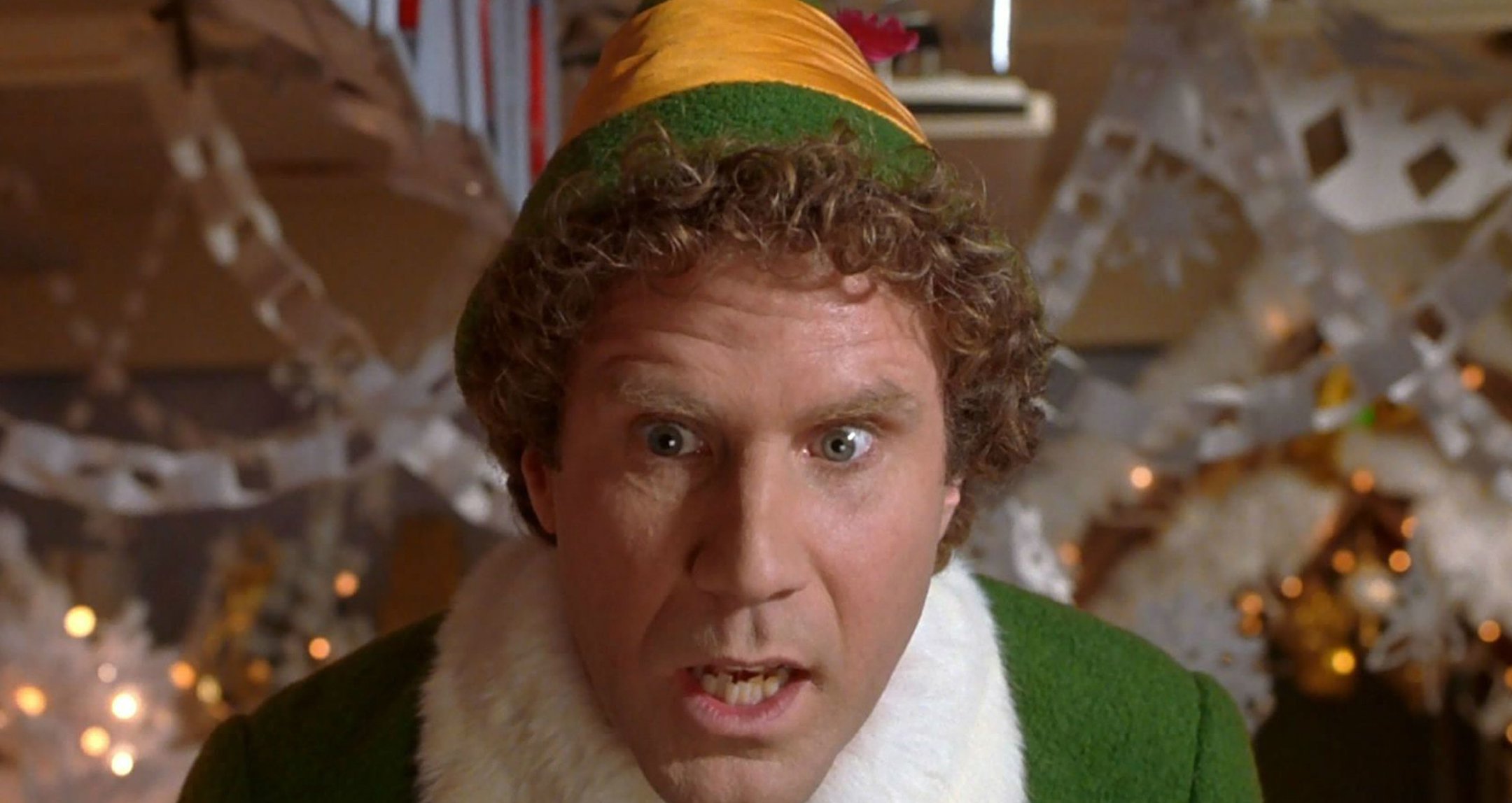 Close up of Will Ferrell in the film Elf, dressed as an elf with a surprised expression, Christmas decorations behind him.