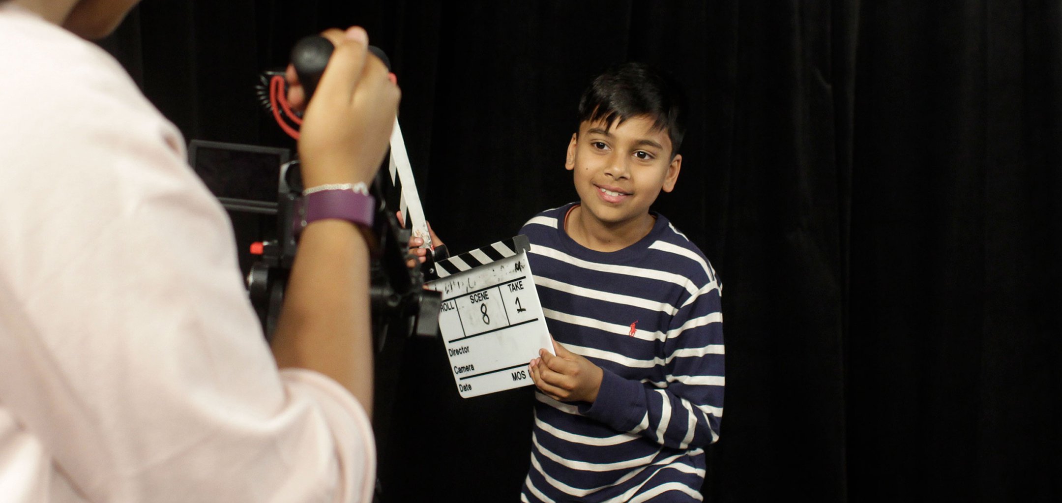 Kid holding a clapperboard in front of a black screen and a video camera